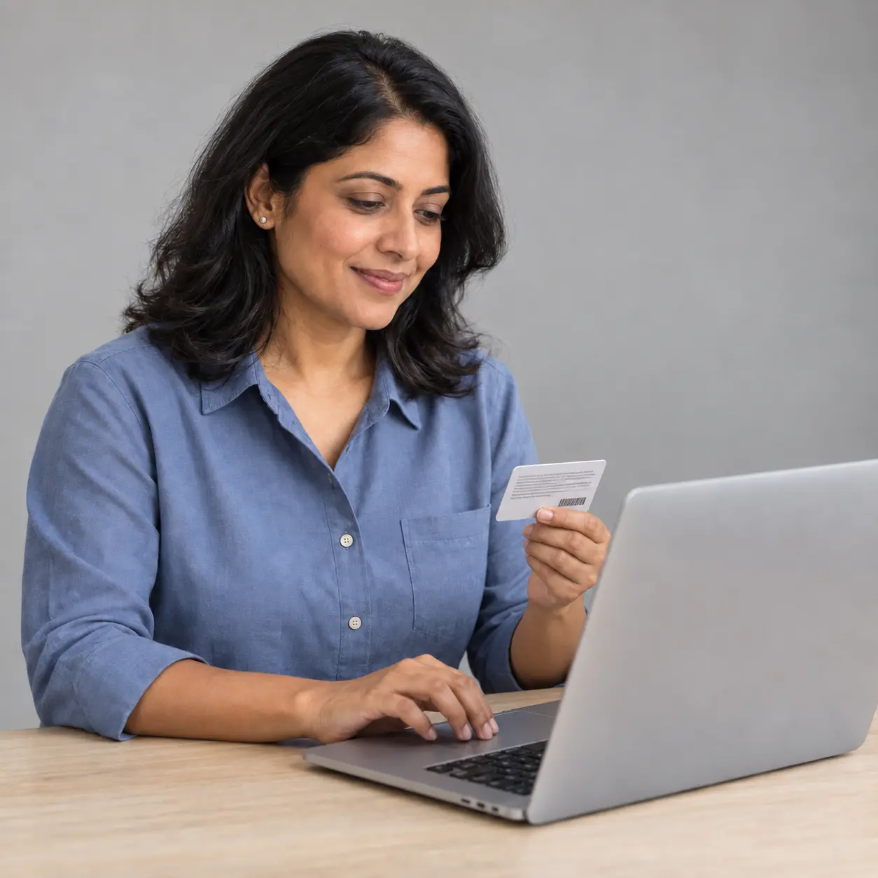 A woman in a blue shirt at a desk, holding her insurance card while she types on a laptop. Verify insurance coverage at Ridge Way.